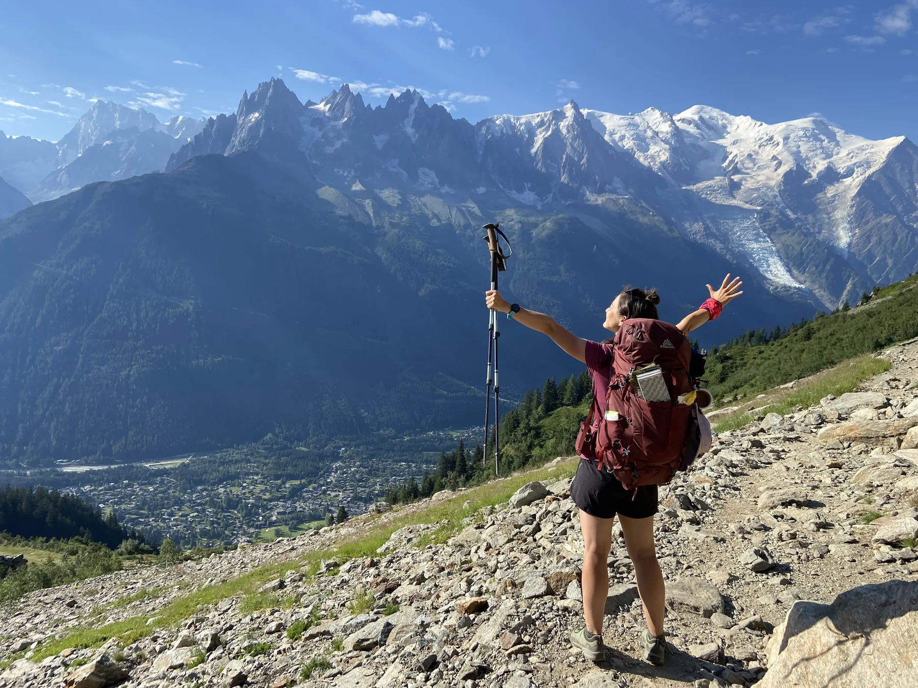 A hiker with arms raised in front of snow-capped Alpine peaks under a blue sky