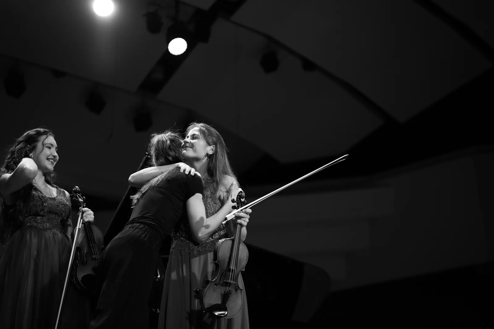 Black-and-white photo of Maria hugging a violinist on stage, with two other violinists smiling beside them